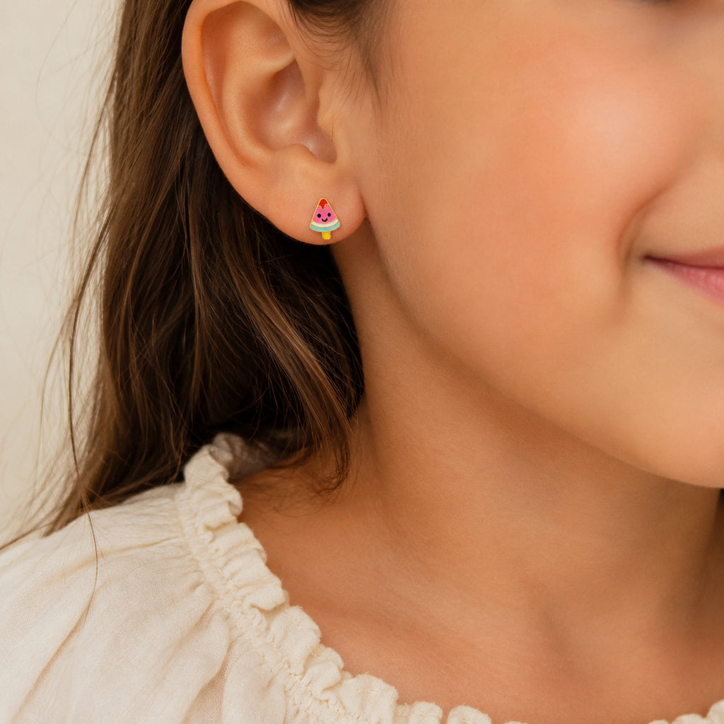 A close-up of a child's ear wearing a small pastel rainbow cupcake stud earring, with the child's face partially visible against a soft light background.