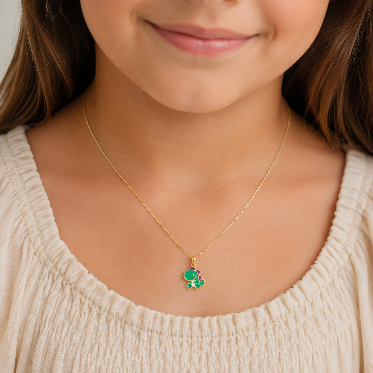 A close-up of a child wearing a delicate gold chain necklace with a small green elephant charm, styled with a cream-colored top.