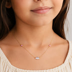 A close-up of a child wearing a delicate gold necklace with a small cloud charm set with clear crystals, flanked by two small pink flower charms, on a light gray background.