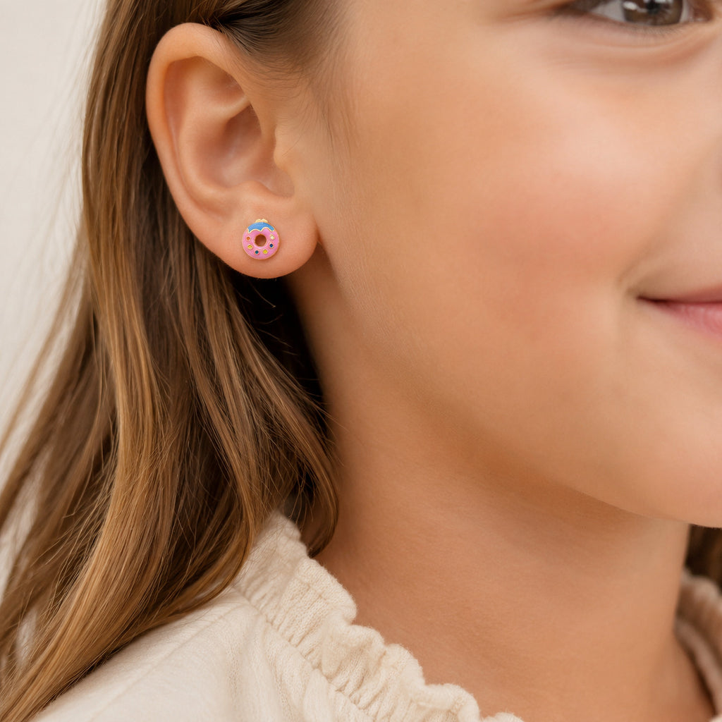 A close-up of a child's ear wearing a small donut-shaped stud earring with pink frosting and colorful sprinkles, against a soft light background.