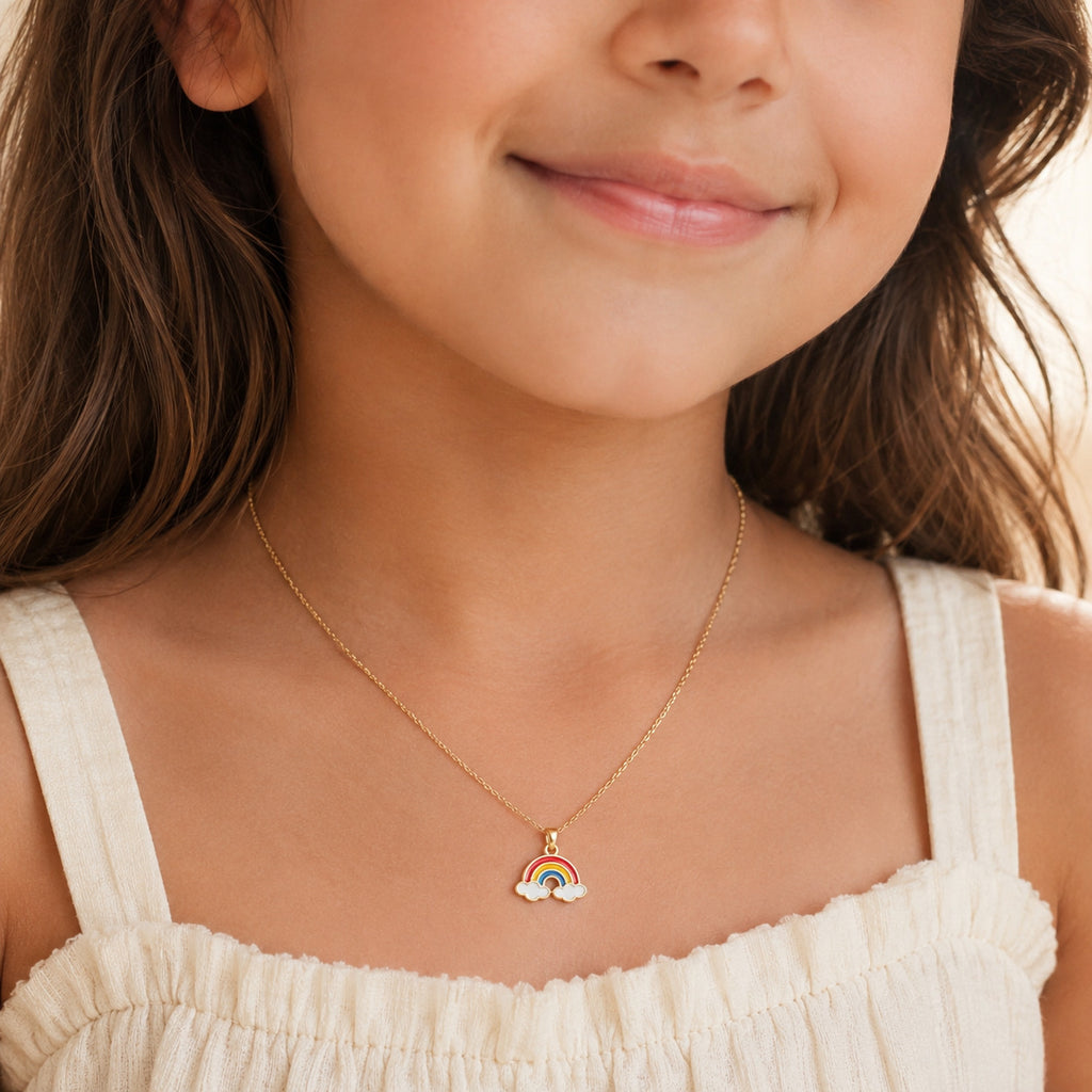 A close-up lifestyle shot of a young girl wearing a delicate gold chain necklace with a small rainbow and cloud pendant, paired with a cream-colored top.