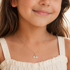 A close-up lifestyle shot of a young girl wearing a delicate gold chain necklace with a small rainbow and cloud pendant, paired with a cream-colored top.