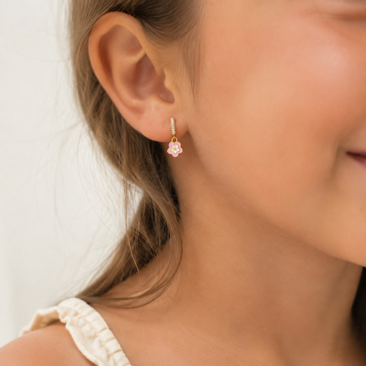 A close-up of a child's ear wearing a small gold dangle earring with a pink and white flower charm.