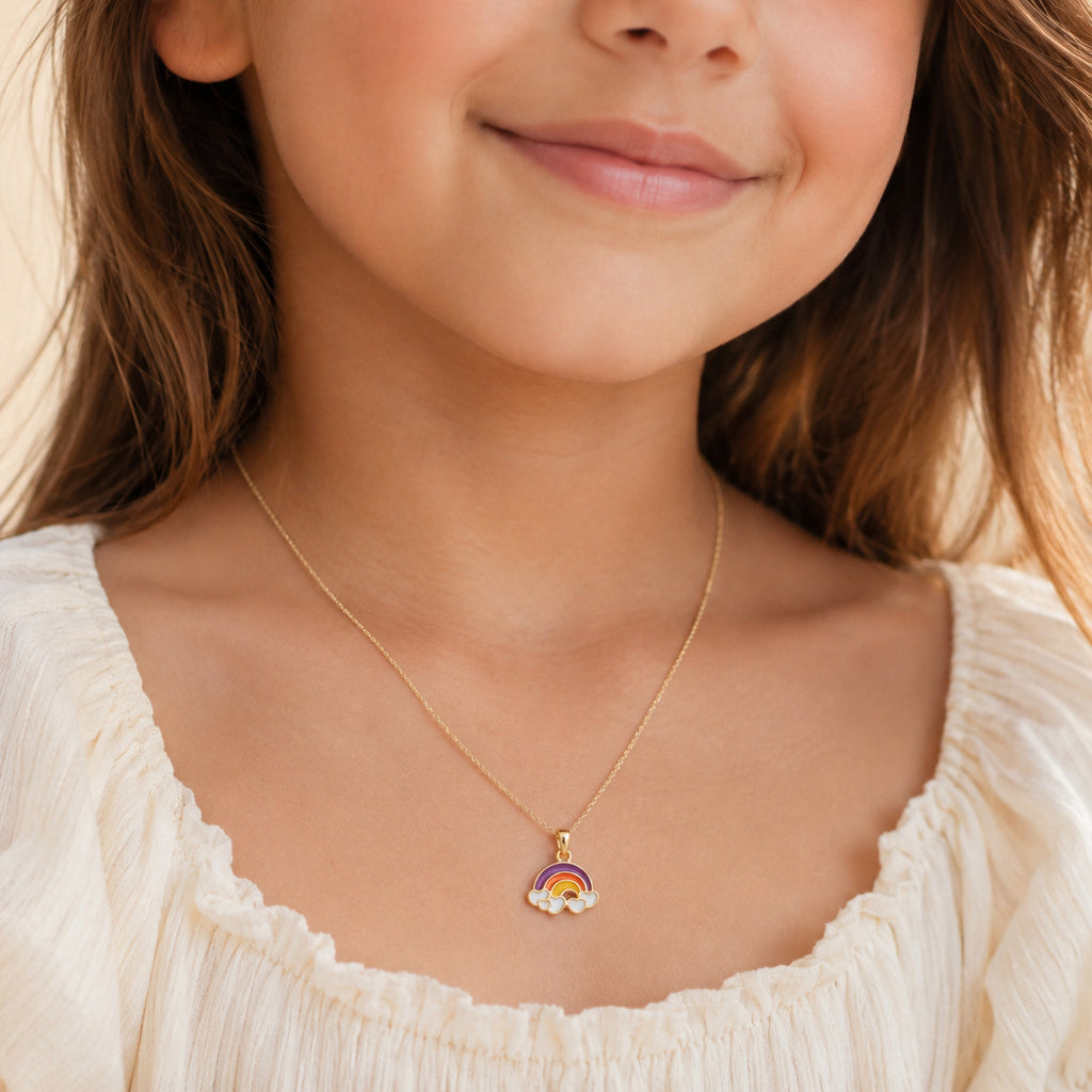 A close-up of a young girl wearing a delicate gold chain necklace with a small rainbow and flower charm, paired with a cream-colored blouse.