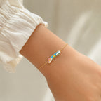 A delicate gold chain bracelet with a small rainbow and cloud charm, worn on a person's wrist. The person is wearing a white ruffled sleeve, and the background is a soft, warm beige.