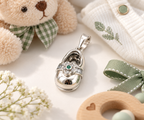 Elegant baby shoe charm pendant with green flower detail displayed beside a teddy bear and soft baby accessories.