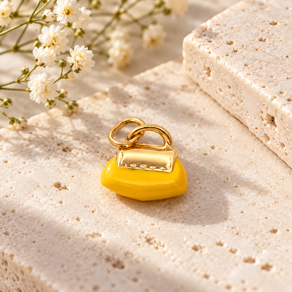 Yellow handbag-shaped jewelry charm with gold-tone details, displayed on natural beige stone beside delicate white flowers.
