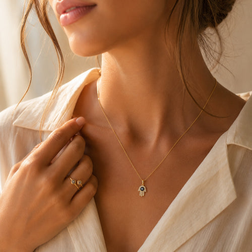 Woman wearing a 14K yellow gold Hamsa evil eye necklace with blue enamel center and CZ stones, styled as an everyday fine jewelry pendant