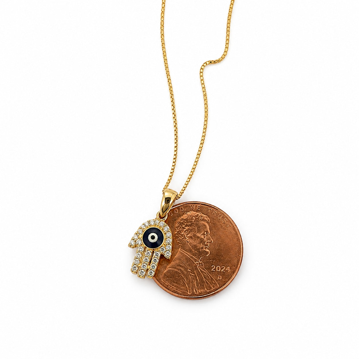 14K yellow gold Hamsa evil eye pendant necklace shown beside a U.S. penny for size reference, featuring blue enamel detailing and sparkling CZ stones
