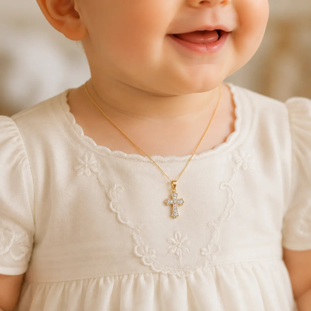 A close-up of a baby wearing a delicate gold cross pendant necklace with clear crystals, styled with a cream-colored embroidered dress.
