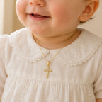 A close-up of a baby wearing a delicate gold cross pendant necklace with small clear stones, paired with a cream-colored embroidered dress.